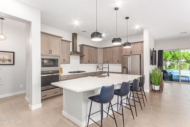 kitchen with pendant lighting, wall chimney exhaust hood, stainless steel appliances, sink, and a kitchen island with sink