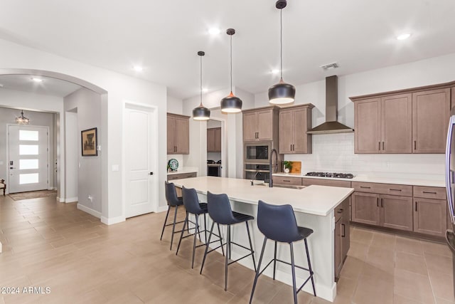 kitchen featuring wall chimney exhaust hood, stainless steel appliances, sink, hanging light fixtures, and a center island with sink