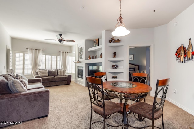 dining room featuring light carpet, baseboards, a glass covered fireplace, ceiling fan, and built in shelves