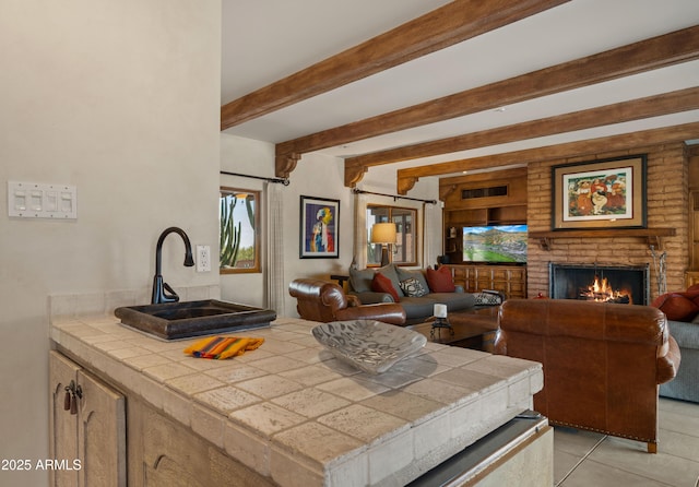 kitchen with beam ceiling, tile counters, a brick fireplace, open floor plan, and a sink