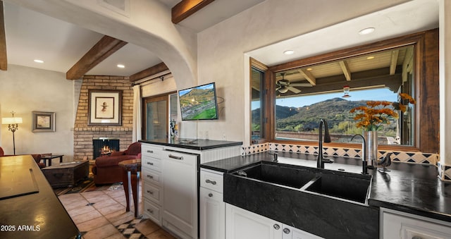 kitchen featuring dark countertops, beamed ceiling, a brick fireplace, white cabinetry, and a sink