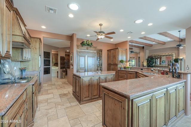 kitchen featuring sink, light stone counters, a center island with sink, kitchen peninsula, and beam ceiling