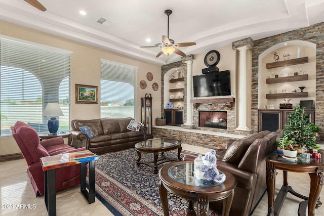 tiled living room with a raised ceiling, a stone fireplace, ceiling fan, built in features, and ornate columns