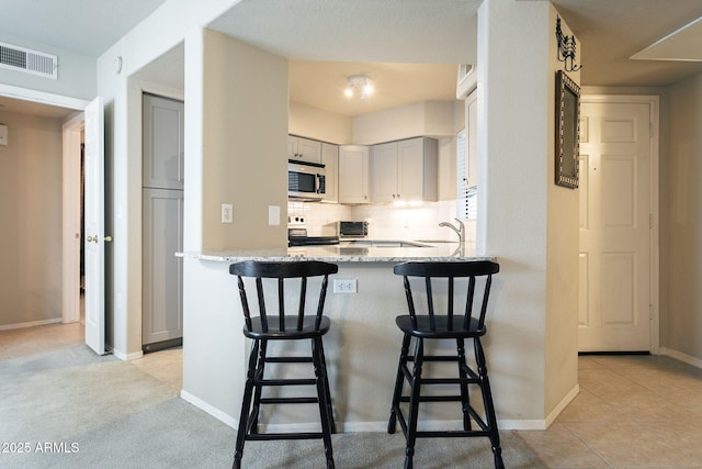 kitchen with kitchen peninsula, a kitchen breakfast bar, backsplash, light stone countertops, and gray cabinetry