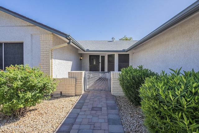 property entrance featuring fence, stucco siding, brick siding, a gate, and a shingled roof