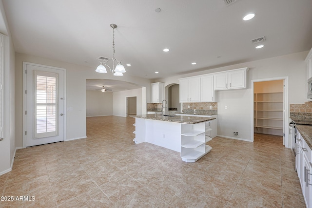 kitchen featuring backsplash, ceiling fan with notable chandelier, stone countertops, a center island with sink, and hanging light fixtures