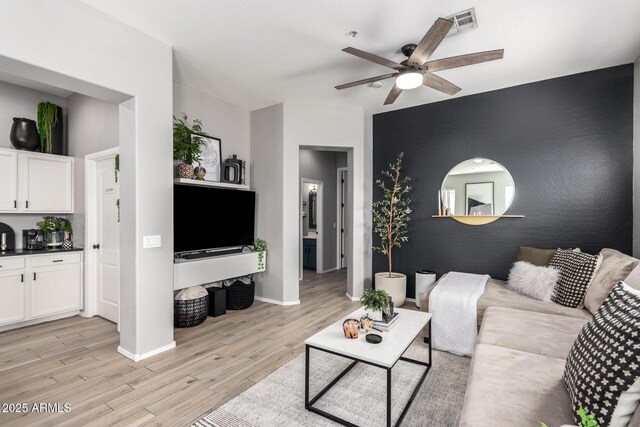 living area with light wood-type flooring, visible vents, baseboards, ceiling fan, and an accent wall