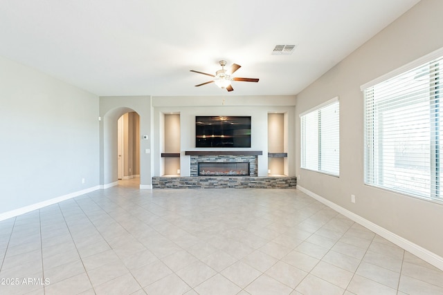 unfurnished living room with arched walkways, visible vents, a ceiling fan, a stone fireplace, and baseboards