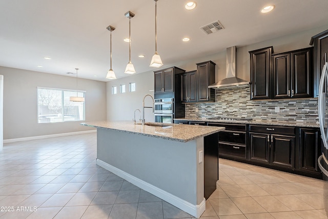 kitchen featuring a kitchen island with sink, stainless steel appliances, visible vents, wall chimney range hood, and decorative light fixtures