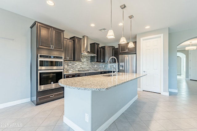 kitchen featuring arched walkways, stainless steel appliances, a sink, wall chimney range hood, and an island with sink