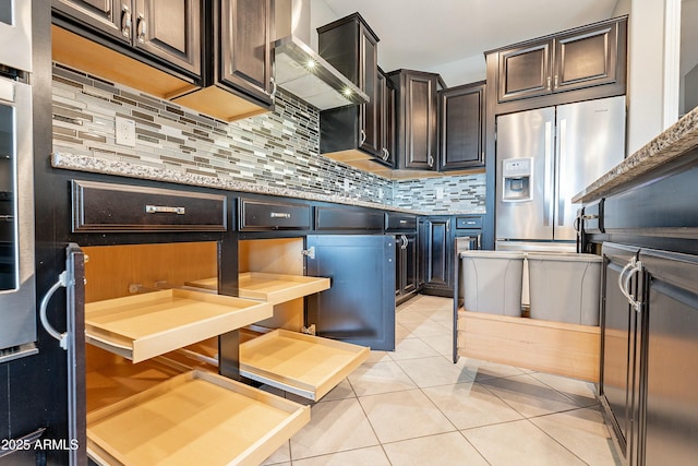 kitchen with dark brown cabinetry, light tile patterned floors, decorative backsplash, light stone counters, and wall chimney range hood