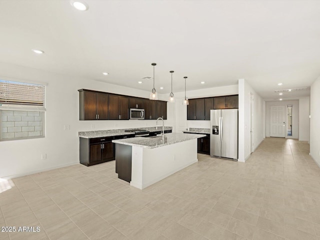 kitchen with hanging light fixtures, stainless steel appliances, dark brown cabinetry, light stone counters, and a center island with sink