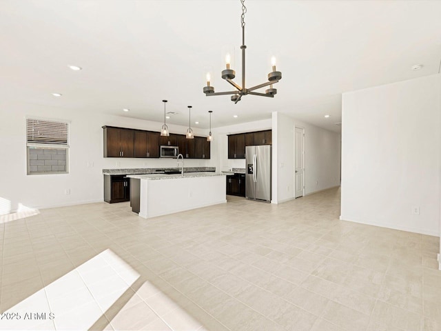 kitchen featuring hanging light fixtures, stainless steel appliances, dark brown cabinetry, light stone countertops, and an island with sink