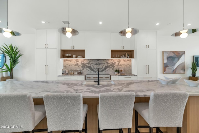 kitchen with a kitchen island with sink, tasteful backsplash, decorative light fixtures, light stone countertops, and white cabinetry