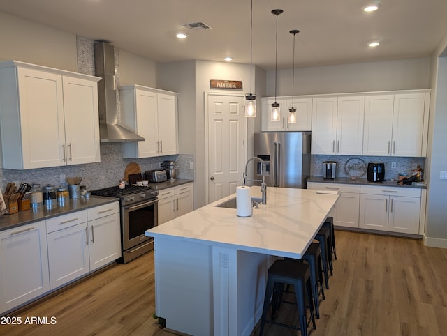 kitchen with high quality appliances, sink, white cabinets, wall chimney range hood, and a center island with sink