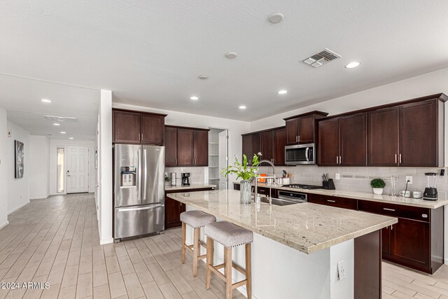 kitchen featuring stainless steel appliances, a center island with sink, light stone counters, a kitchen breakfast bar, and backsplash