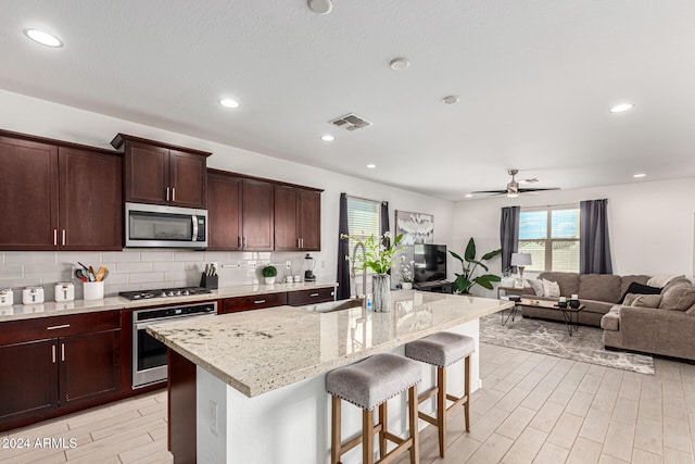 kitchen with stainless steel appliances, light stone countertops, a breakfast bar area, ceiling fan, and a kitchen island with sink