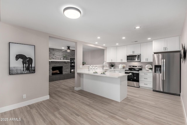 kitchen with a stone fireplace, appliances with stainless steel finishes, white cabinets, light wood-type flooring, and kitchen peninsula