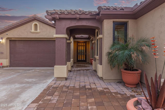 exterior entry at dusk featuring a tiled roof, an attached garage, driveway, and stucco siding