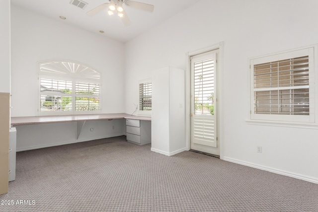 unfurnished office with ceiling fan, visible vents, built in desk, baseboards, and carpet flooring