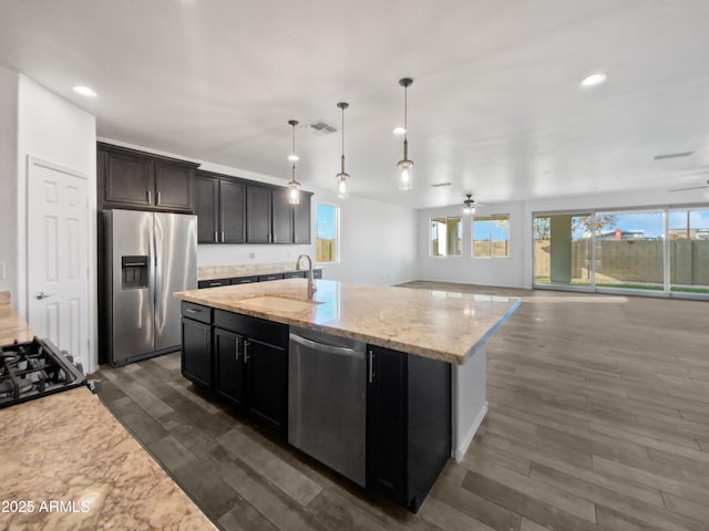 kitchen with sink, appliances with stainless steel finishes, hanging light fixtures, light stone counters, and a center island with sink