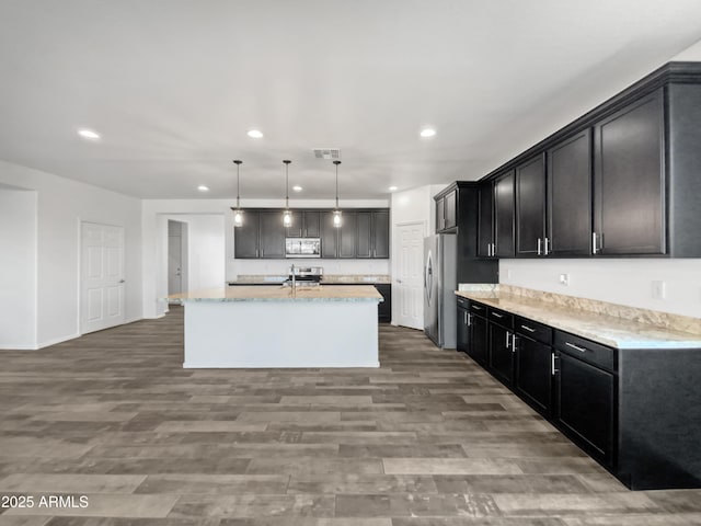 kitchen with appliances with stainless steel finishes, decorative light fixtures, an island with sink, wood-type flooring, and light stone counters