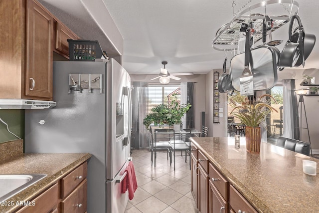 kitchen with brown cabinetry, light tile patterned floors, under cabinet range hood, and stainless steel refrigerator with ice dispenser