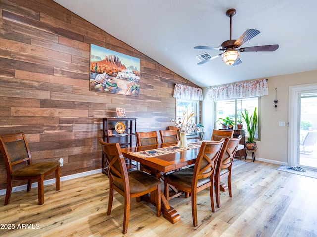 dining area with a ceiling fan, light wood-style flooring, wood walls, lofted ceiling, and visible vents