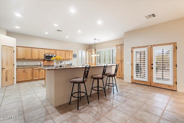 kitchen featuring light brown cabinets, an island with sink, a kitchen breakfast bar, light stone countertops, and pendant lighting