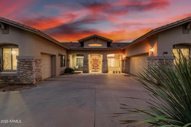 exterior entry at dusk featuring concrete driveway, a garage, stone siding, and stucco siding