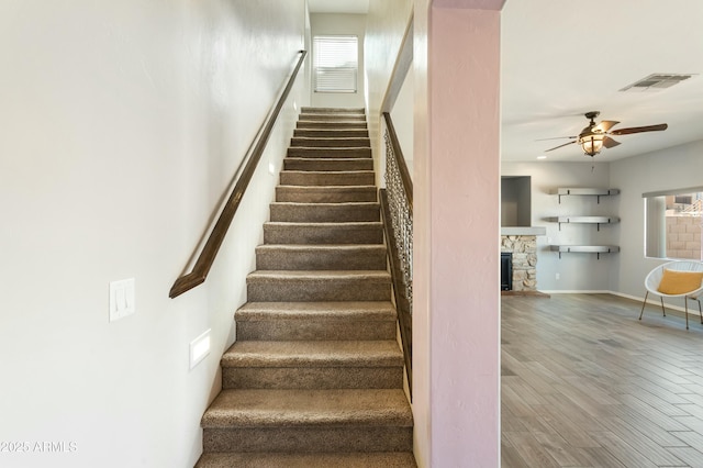 stairs with ceiling fan, wood-type flooring, and a stone fireplace