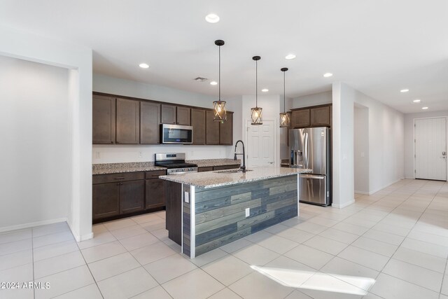 kitchen featuring hanging light fixtures, an island with sink, appliances with stainless steel finishes, light stone countertops, and dark brown cabinetry