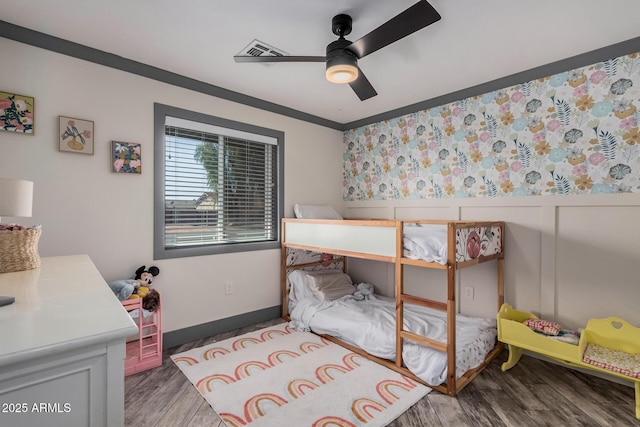 bedroom featuring ornamental molding, a ceiling fan, wood finished floors, wainscoting, and wallpapered walls