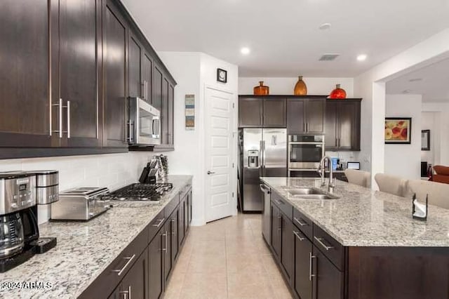 kitchen with light stone counters, an island with sink, light tile patterned floors, sink, and stainless steel appliances