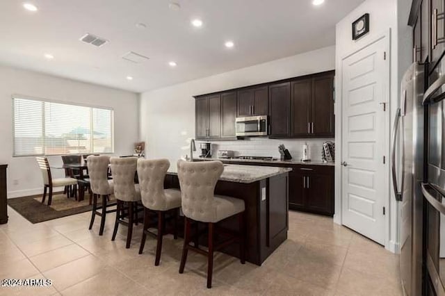 kitchen with a kitchen breakfast bar, stainless steel appliances, light stone countertops, an island with sink, and light tile patterned floors