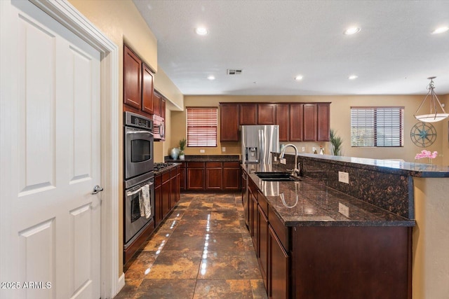 kitchen with sink, hanging light fixtures, dark stone countertops, an island with sink, and appliances with stainless steel finishes