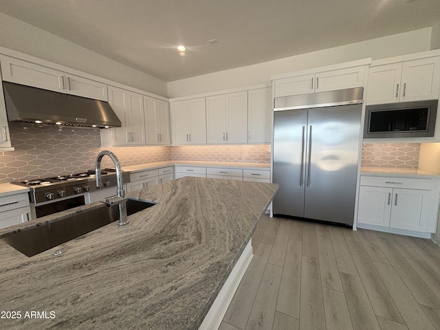 kitchen with built in appliances, under cabinet range hood, backsplash, and white cabinets