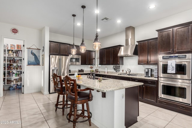 kitchen featuring decorative light fixtures, a kitchen island with sink, stainless steel appliances, light stone countertops, and wall chimney exhaust hood