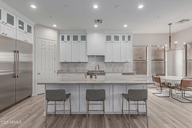 kitchen featuring wall chimney exhaust hood, light stone counters, white cabinetry, built in refrigerator, and an island with sink