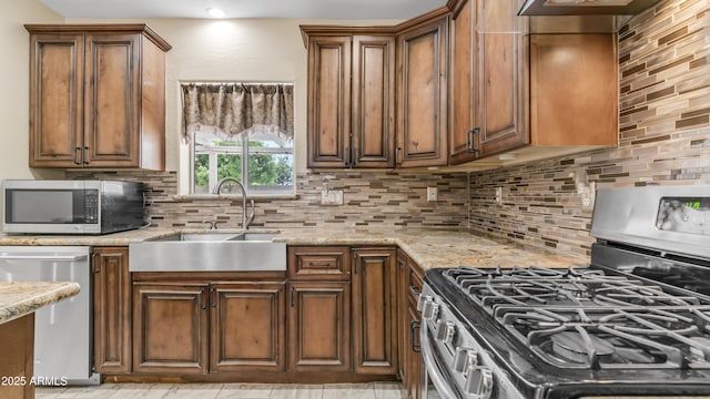 kitchen featuring stainless steel appliances, a sink, light stone countertops, decorative backsplash, and brown cabinetry