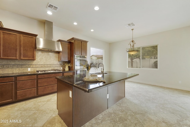 kitchen with sink, dark stone countertops, decorative backsplash, wall chimney range hood, and a center island with sink