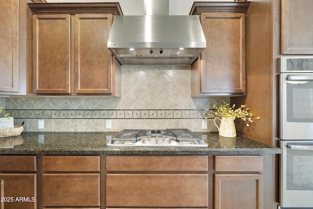 kitchen with dark stone countertops, stainless steel appliances, tasteful backsplash, and wall chimney range hood