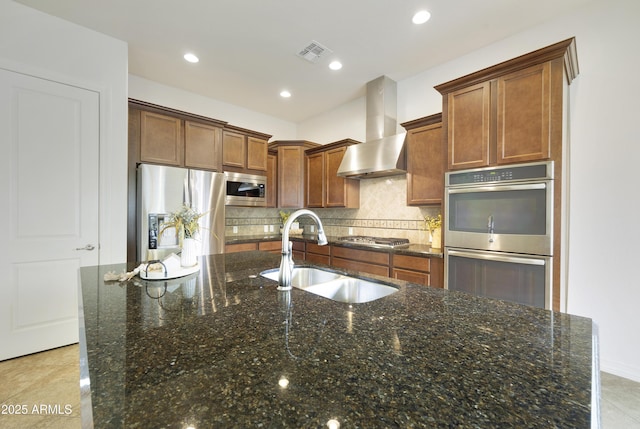 kitchen with sink, a center island with sink, appliances with stainless steel finishes, dark stone counters, and wall chimney range hood