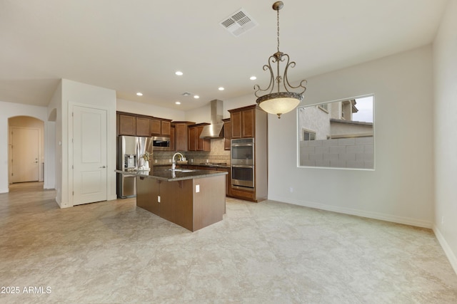 kitchen with appliances with stainless steel finishes, pendant lighting, an island with sink, dark stone counters, and wall chimney range hood