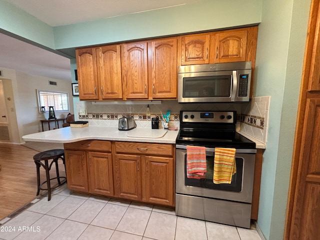 kitchen with light tile patterned flooring, backsplash, appliances with stainless steel finishes, a kitchen bar, and kitchen peninsula