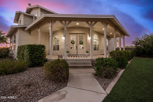 exterior entry at dusk featuring a porch, french doors, stone siding, and a lawn