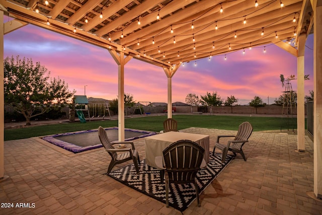 patio terrace at dusk featuring outdoor dining space, a playground, a lawn, and fence