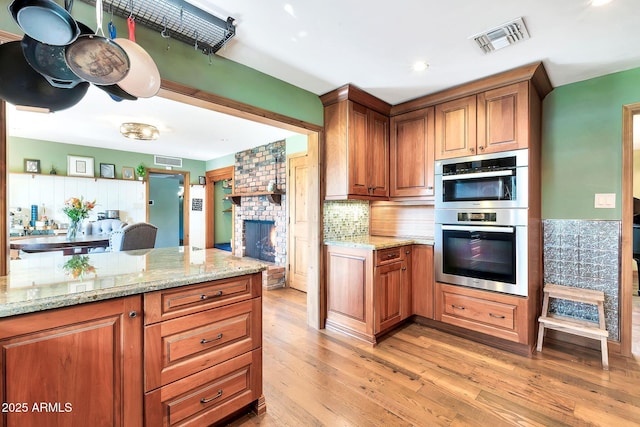 kitchen featuring double oven, decorative backsplash, light stone countertops, a brick fireplace, and light wood-type flooring