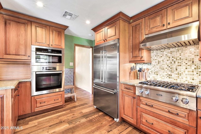 kitchen featuring range hood, tasteful backsplash, wood-type flooring, light stone counters, and stainless steel appliances