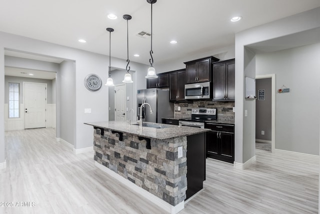 kitchen featuring decorative light fixtures, backsplash, a kitchen island with sink, stainless steel appliances, and light stone counters